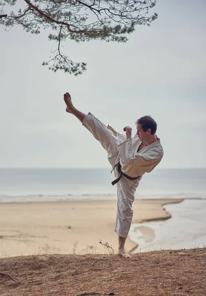 Karate man in an old kimono and black belt training high kick at the sea. Martial arts concept. — Stock Photo, Image
