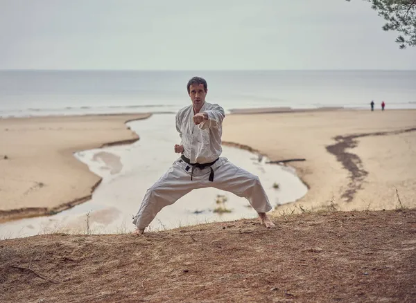 Karate man in an old kimono and black belt training punch at the sea. Martial arts concept. — Stock Photo, Image