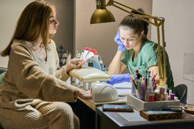 Müşteri, manikür için gösterilen verniğin kırmızı tonunu seçer. Mavi eldivenli usta bekliyor..