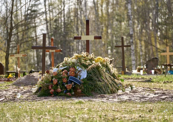 New grave with wooden cross and several funeral wreaths. Another graves with crosses behind it. Forest at the background.