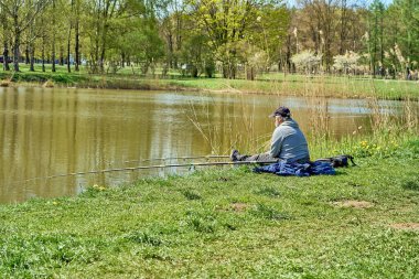 Riga, Letonya - 8 Mayıs 2022: Bir balıkçı halka açık bir parktaki bir gölün kıyısında elinde olta ile oturur. Etrafta yeşil çimenler var. Arkadan görüntüle.
