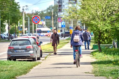 RIGA, LATVIA - MAY 13, 2022: Woman riding bicycle on a sidewalk. A man in a coat and sunglasses walks in the opposite direction during the sunny day.