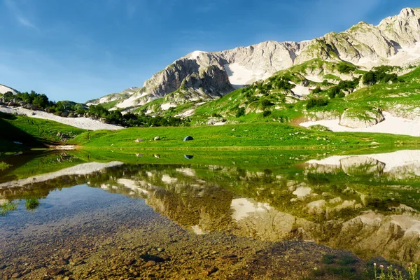 Zeltlager am grün blühenden Ufer eines von Berggipfeln umgebenen Bergsees lizenzfreie Stockfotos