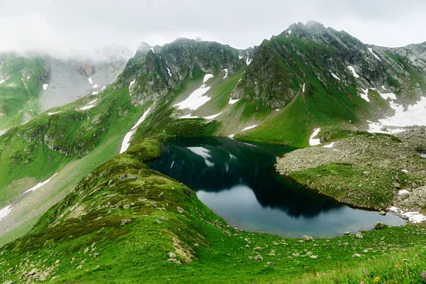 Berglandschaft mit kleinem Gletschersee. Berggipfel mit Schnee, im Vordergrund grünes Gras Stockbild