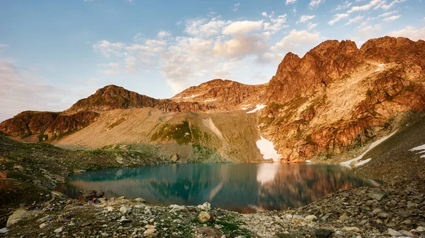 Paesaggio con alte rocce cime illuminate, pietre nel lago di montagna, cielo nuvoloso blu e luce solare gialla all'alba — Foto Stock