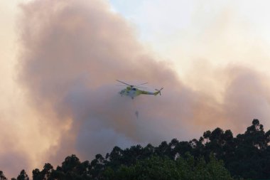 Bir helikopter orman yangınından çıkan duman bulutlarının arasında su boşaltarak uçar.