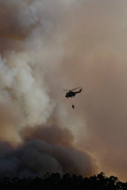 Bir helikopter orman yangınından çıkan duman bulutlarının arasında su boşaltarak uçar.