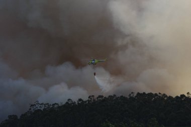 Bir helikopter orman yangınından çıkan duman bulutlarının arasında su boşaltarak uçar.