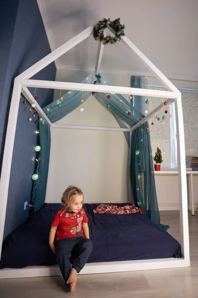 可爱的孩子对她房间里的圣诞装饰印象深刻.女孩穿着红T恤坐在床上 — 图库照片