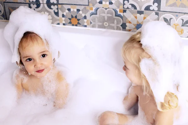 Две маленькие девочки-сестры веселятся в ванной, играя с пеной для ванны. Уход за детьми — стоковое фото