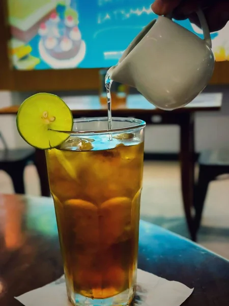 liquid sugar is poured into a glass of iced tea with lemon slices on a wooden table in a cafe on a blurred background