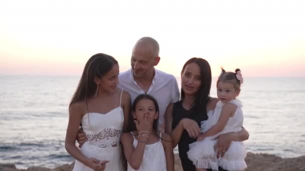 Wielopokoleniowa, kaukaska rodzina wysyłająca pocałunki w zwolnionym tempie patrząc na kamerę uśmiechniętą o zachodzie słońca na wybrzeżu morza. Pozytywny szczęśliwy ojciec matka i córki w śródziemnomorskim krajobrazie. — Wideo stockowe