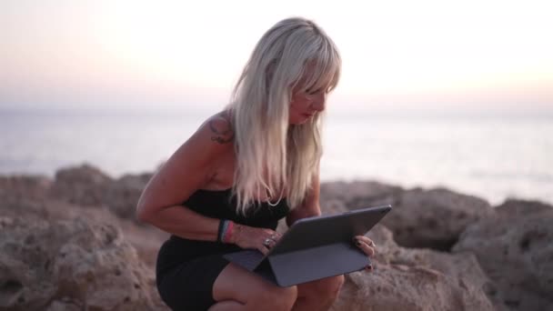 Αυτοπεποίθηση Καυκάσια ώριμη γυναίκα κάθεται σε βράχους στη Μεσόγειο θάλασσα θαυμάζοντας το ηλιοβασίλεμα μηνυμάτων σε απευθείας σύνδεση στο tablet. Πορτρέτο της αυτοπεποίθηση επιτυχημένη επιχειρηματίας που εργάζονται σε απευθείας σύνδεση για διακοπές σε εξωτερικούς χώρους. — Αρχείο Βίντεο