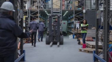 Kapalı alanda çalışan insanlarla dolu geniş açılı bir depo. Kendine güvenen forklift operatörü yavaş çekimde depo otomatik yükleyicisini kullanıyor..