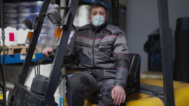 Portret van een blanke volwassen man in een coronavirus masker zittend in een vorkheftruck kijkend naar de camera. Zelfverzekerde serieuze werknemer in uniform poseren in autolader in industriële opslag op Covid-19 pandemie. — Stockvideo