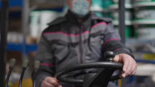 Close-up-Gabelstaplerlenkrad mit männlichen kaukasischen Händen darauf. Unscharfer, unkenntlich gemachter Fahrer mit Covid-19-Gesichtsmaske sitzt in Maschinen im Hintergrund in der industriellen Lagerung der Coronavirus-Pandemie. — Stockvideo