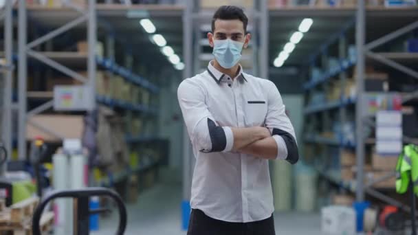 Fiducioso uomo mediorientale in maschera facciale Covid mani incrociate guardando la fotocamera in piedi in magazzino industriale al chiuso. Ritratto di orgoglioso manager professionista in posa sul posto di lavoro in caso di pandemia. — Video Stock