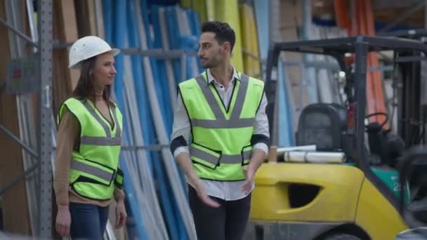 Hombre de Oriente Medio y mujer caucásica caminando en el almacén hablando. Cámara en vivo sigue a los empleados que planifican el envío de carga en interiores en almacenamiento industrial discutiendo los plazos logísticos. — Vídeo de stock