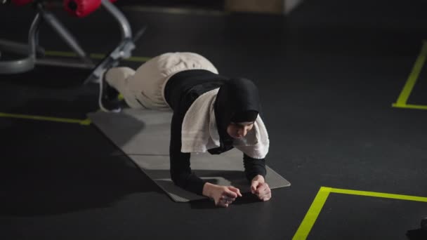 Szeroki strzał wytrzymała kobieta z Bliskiego Wschodu stojąca na desce i siedząca na macie do ćwiczeń. Pasuje szczupła sportsmenka w hidżabie ćwicząca na siłowni w domu. Zdrowy styl życia i koncepcja fitness. — Wideo stockowe