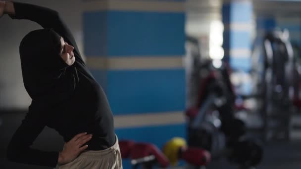 Mujer motivada feliz en hijab calentando los músculos estirando las manos y la cintura de pie en el interior del gimnasio. Deportista de confianza en forma de slim Oriente Medio haciendo ejercicio. Concepto deportivo. — Vídeos de Stock