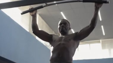 Afrikalı Amerikalı sporcu, kaslı vücudunda güneş ışığıyla barfiks çekiyor. Barda dinlenen güçlü motive edilmiş, üstsüz bir adamın orta boy fotoğrafı. Canlı kamera hareket ediyor.