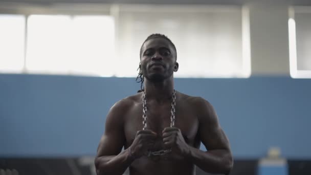 Retrato de confiante sério afro-americano sem camisa homem posando no ginásio com corrente no pescoço. médio tiro de forte muscular bonito desportista alongamento de metal com as mãos olhando para a câmera. — Vídeo de Stock