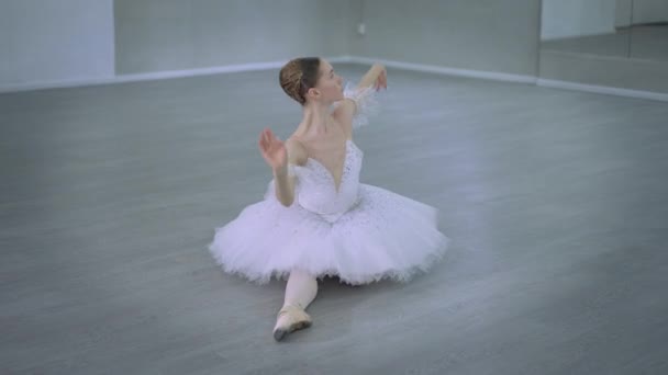 Underbar kvinna i vit tutu klänning i garn sitter på golvet lutande sig ner. Brett filmporträtt av seriös koncentrerad kaukasisk ung ballerina som repeterar i dansstudio inomhus. Konstbegrepp. — Stockvideo