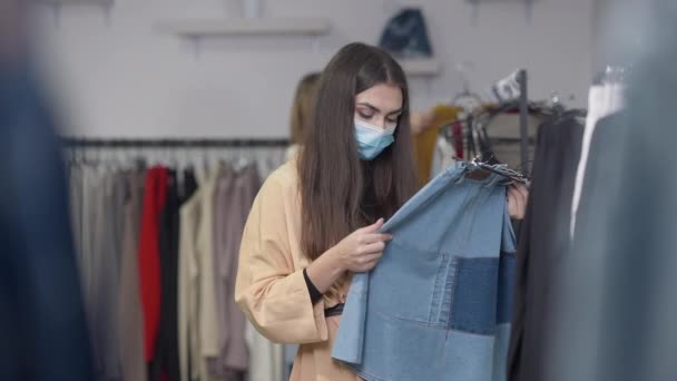 Portret van een geconcentreerde vrouw in coronavirus gezichtsmasker die jeans rok controleert in de winkel op Black Friday verkoop als klant die op de achtergrond passeert. Mooi vertrouwen Kaukasische shopper kiezen aankoop. — Stockvideo