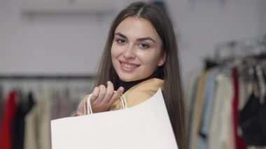 Alışveriş poşetleriyle dükkânda kameraya bakıp gülümseyen muhteşem bir kadın. İçeride poz veren, kendinden emin, beyaz, güzel kadın alıcının portresi. Tüketim kavramı.
