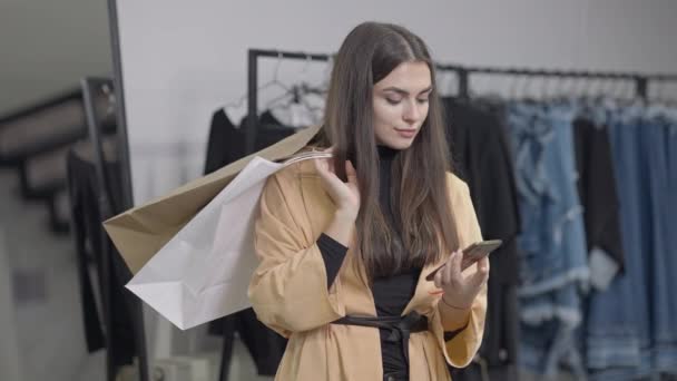 Foto media de mujer rica y segura de sí misma con bolsas de compras de pie en la tienda mirando a la cámara sonriendo y mirando hacia otro lado. Retrato de cliente caucásico rico satisfecho en tienda de ropa con compras. — Vídeo de stock