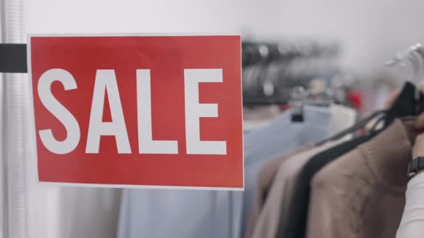 Mensaje de venta en rack con mano caucásica femenina que toma muchas perchas al salir del fondo. Mujer irreconocible de compras eligiendo ropa en la tienda al por menor en interiores en las ventas del Viernes Negro. — Vídeos de Stock