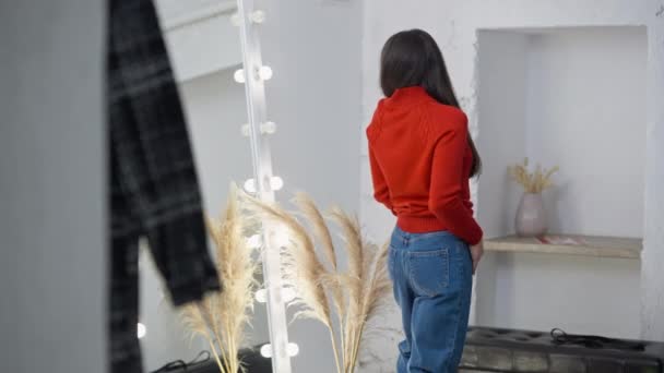 Jovem mulher magro olhando para reflexão experimentando camisola vermelha e jeans jeans jeans azul na loja. Vista lateral do concentrado belo comprador caucasiano em pé na frente do espelho examinando roupa. — Vídeo de Stock