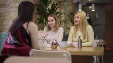 Kafede oturup gülen üç neşeli genç kadın. Güzel, Kafkasyalı ve Orta Doğulu arkadaşlar kahve içmek için içeride sohbet ediyorlar. Yavaş çekim.