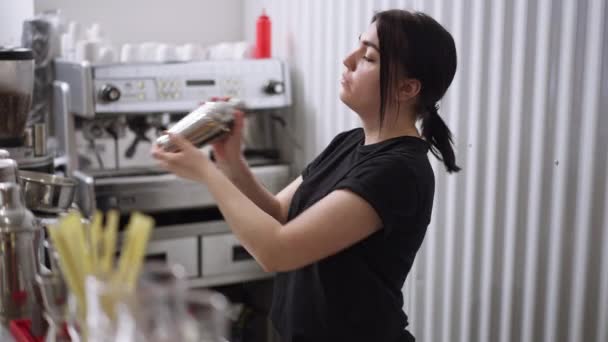 Vista lateral concentrada profesional hembra camarera agitando coctelera en cámara lenta preparando bebida alcohólica. Retrato plano mediano de una joven caucásica delgada enfocada que trabaja en un bar bar. — Vídeo de stock