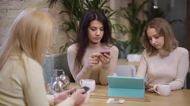 Τρεις νεαρές απορροφημένες γυναίκες της χιλιετίας σερφάρουν στα μέσα κοινωνικής δικτύωσης σε smartphones που κάθονται στο τραπέζι στο εστιατόριο. Απεχθής Μέση Ανατολή και Καυκάσιοι φίλοι αγνοούν ο ένας τον άλλο περιμένοντας την τάξη στο καφέ. — Αρχείο Βίντεο