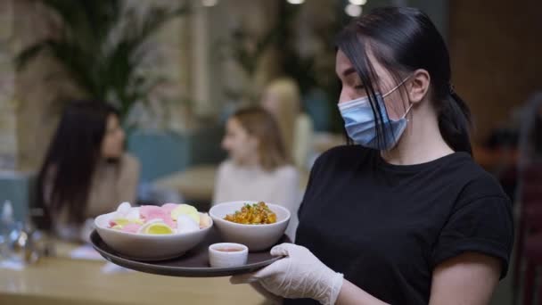 Zelfverzekerde vrouwelijke ober in coronavirus gezichtsmasker poseren met zeevruchten op dienblad en wazig klanten zitten in restaurant op de achtergrond. jonge blanke vrouw kijken naar camera staan met orde. — Stockvideo