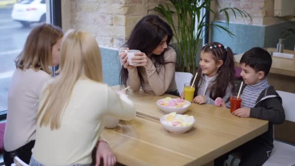 Młoda matka i syn z Bliskiego Wschodu siedzą w kawiarni z białymi kobietami i rozmawiają o piciu. Relaks pięknych przyjaciół i dzieci korzystających z kolacji w restauracji w zwolnionym tempie. — Wideo stockowe