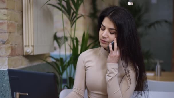 Mujer hermosa ocupada concentrada hablando por teléfono sentado en la cafetería con el ordenador portátil. Retrato de la hermosa mujer de negocios seria y confiada de Oriente Medio discutiendo administración. Éxito y estilo de vida. — Vídeos de Stock