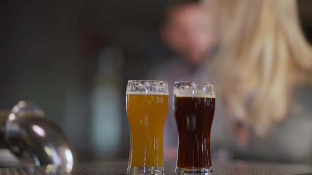 Verres à bière avec lager pâle et sombre sur le comptoir du bar et le couple caucasien flou entrant assis en parlant. Homme et femme méconnaissables sortir ensemble bavarder dans un pub à l "intérieur. Mode de vie et relations — Video