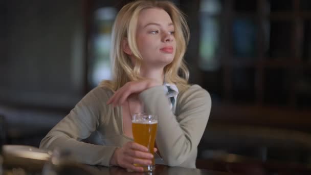 Великолепная стройная белая молодая женщина, сидящая за барной стойкой со стаканом бледного пива. Портрет очаровательной блондинки, ожидающей свидания в пабе в помещении. Уверенность и стиль жизни. — стоковое видео