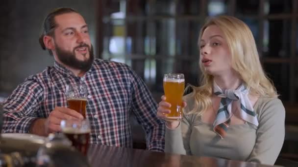 Biała para gadająca brzęczącymi okularami, pijąca piwo w pubie. Piękna młoda kobieta i przystojny brodaty mężczyzna randkujący w barze. Relaks i relacja. — Wideo stockowe