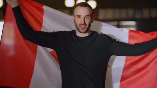 Vooraanzicht jonge knappe blanke man met Canadese vlag die glimlacht terwijl hij naar de camera kijkt. Portret van een zelfverzekerde gelukkige man poseren met nationaal symbool verheugen sportteam succes op kampioenschap. — Stockvideo