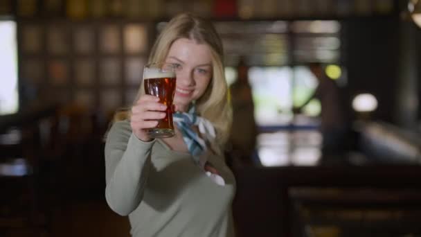 Glad ung kvinna rostning sträcker öl glas till kameran ler stående på puben inomhus. Rack fokus från leende ansikte till pint av lager och tillbaka. Helg fritid och livsstil koncept. — Stockvideo