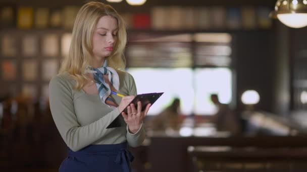 Jeune femme concentrée commande par écrit avec stylo servant les clients dans le restaurant à l'intérieur. Portrait d'une serveuse caucasienne mince et charmante travaillant dans un pub. Profession et professionnalisme. — Video