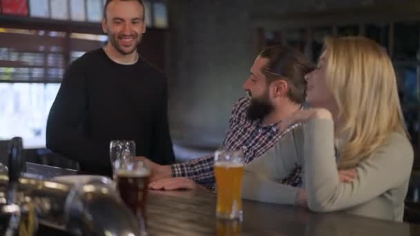 Joyeux couple caucasien saluant ami entrant bar assis au comptoir boire de la bière. Positif heureux détendus hommes et femmes se réunissant dans un pub reposant le week-end. Mode de vie et amitié. — Video