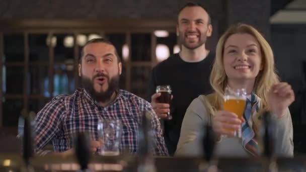 Excited Caucasian friends rejoicing goal watching sport match in pub indoors. Portrait of happy smiling men and woman talking smiling resting on weekend at bar counter with beer glasses. — Stock Video