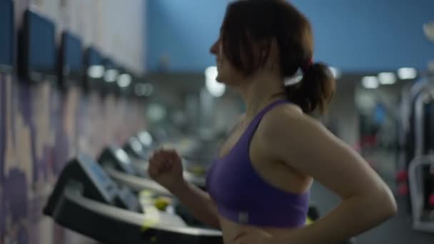 Tampilan samping tersenyum wanita muda Kaukasia ramping joging di treadmill di dalam ruangan olahraga. Happy dimotivasi fit olahragawan wanita berjalan pada peralatan kardio menikmati gaya hidup sehat dan kebugaran. Konsep olahraga. — Stok Video