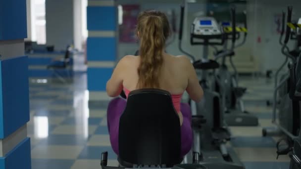 Вид сзади: мотивированная женщина plus-size на велосипеде в тренажерном зале в помещении. Молодая кавказская спортсменка с избыточным весом тренируется, теряя вес. Здоровый образ жизни и мотивация. — стоковое видео