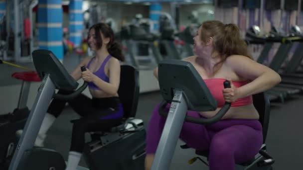 Glad plus storlek vit kvinna cykling motionscykel med smal ung dam tränar i bakgrunden. Två positiva vänner som har roligt tävlar i hastighet på gym utrustning inomhus. Idrottsbegreppet. — Stockvideo