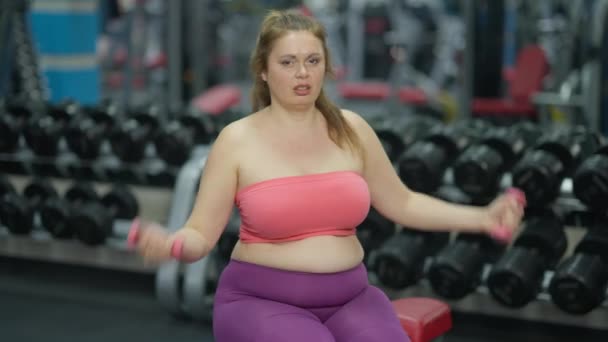 Stolt självsäker plus-size kvinna lyfta hantlar sitter på träningsbänk i gym. Porträtt av ihållande motiverade kaukasiska dam tränar inomhus muskler. Bodybuilding och sport koncept — Stockvideo